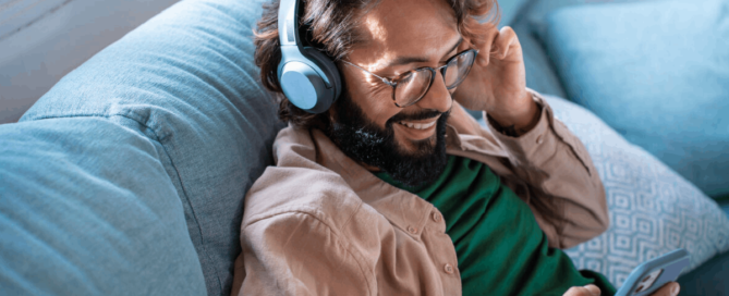 A man wearing headphones and smiling while listening to music on his phone as he relaxes on a couch.